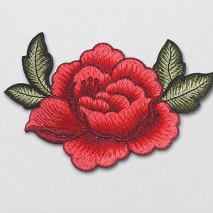 May include: An embroidered patch featuring a large, vibrant red rose with layered petals and detailed stitching. The flower is flanked by two dark green leaves on each side, showcasing intricate thread work and a defined, scalloped edge design.