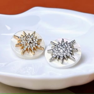 May include: Two decorative buttons featuring a starburst design. One button has a gold-tone starburst with clear crystal accents, the other a silver-tone starburst with clear crystal accents. Both are set on a white, round, mother-of-pearl base.