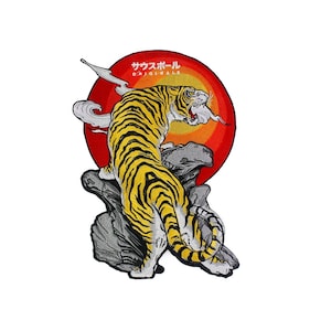 Può includere: Una toppa ricamata con un tigre giallo e nero in piedi su una roccia con un sole rosso e arancione alle sue spalle. Il testo "サウスポール ORIGINALS" è sopra il tigre.