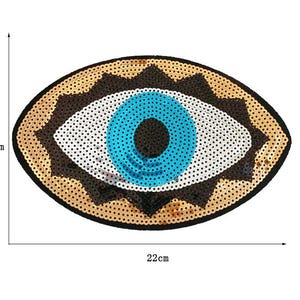 May include: An oval-shaped sequined patch in the shape of an eye. The eye features a blue iris, white sclera, and a brown and gold sequined border. The patch measures approximately 9 inches wide and 5.5 inches tall.