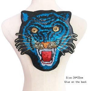 Könnte beinhalten: Ein paillettenbesetzter Tigerkopf-Aufnäher in Blau-, Schwarz-, Gold-, Orange- und Rottönen. Der Aufnäher ist ca. 24 x 22 cm groß und zum Aufkleben bestimmt. Das Maul des Tigers ist geöffnet und zeigt eine rote Zunge.