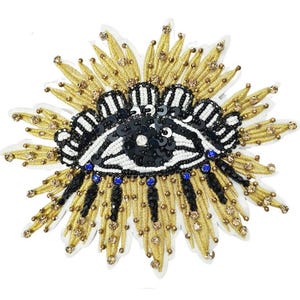 Könnte beinhalten: Gesticktes Augen-Patch mit goldenem, schwarzem und weißem Design. Der Patch zeigt ein zentrales Augenmotiv mit schwarzen Pailletten, weißen Perlen und blauen Akzenten, umgeben von goldfarbenen Verzierungen.