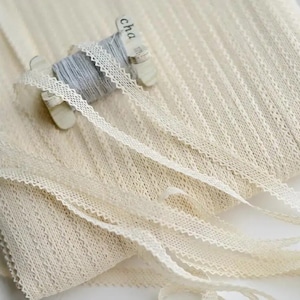 Puede incluir: Una bobina de encaje blanco marfil con un borde festoneado. El encaje está enrollado alrededor de una bobina de cartón con la palabra "Cha" impresa en ella.