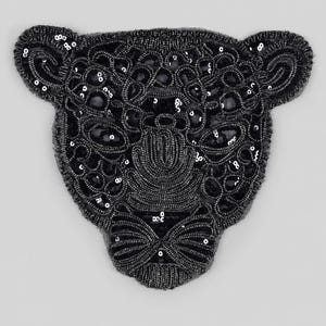May include: A black beaded patch in the shape of a leopard head. The design features intricate embroidery with black glass beads and small circular sequins, creating a textured, detailed pattern that outlines the facial features of the animal.