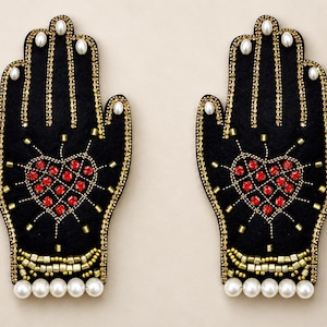 May include: A pair of black fabric hand-shaped patches featuring embroidered red heart motifs, gold bead accents, and white pearl-like embellishments along the fingers and wrists. The design includes intricate gold stitching and red crystals.