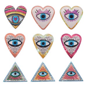 May include: Nine colorful embroidered patches featuring an evil eye design. The patches are in the shapes of hearts and triangles. The colors include pink, blue, yellow, and white.