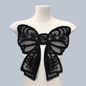 Könnte beinhalten: Eine große schwarze Spitzen-Schleifen-Applikation mit einem zarten Blumenmuster. Die Schleife besteht aus einem durchsichtigen Stoff mit einer strukturierten Oberfläche.