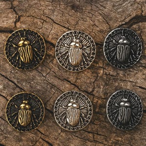 May include: Six metallic round buttons with a scarab beetle design. The buttons are in gold, silver, and gunmetal finishes, each with intricate details. They are arranged on a textured, dark wood surface.