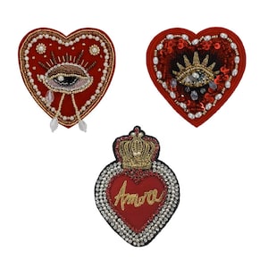 May include: Three heart-shaped embroidered patches. The patches are red, with intricate designs including eyes, crowns, and the word "Amore". Embellishments include beads, sequins, and gold thread, creating a detailed and ornate appearance.