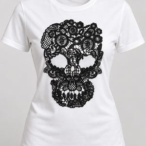 May include: A white short-sleeved t-shirt featuring a large, intricate black lace appliqué in the shape of a human skull centred on the front. The lace design incorporates various floral and geometric patterns, creating a detailed texture.