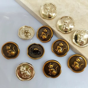 May include: Assortment of 10 gold and bronze cat buttons. The buttons are round and have a raised design of a cat sitting. The buttons are approximately 2.5 cm in diameter.