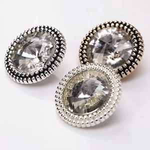 Op de afbeelding: Drie ronde, decoratieve knopen met een zilveren en gouden afwerking. Elke knop heeft een helder, facetgeslepen kristal in het midden.