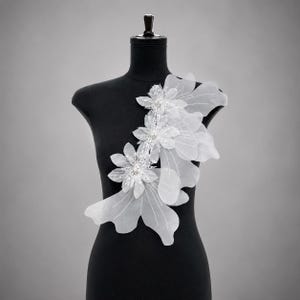 May include: A white floral applique featuring layered sheer fabric petals and intricate beaded centers. The decorative piece is arranged in a vertical cluster of three distinct flower shapes with delicate thread detailing on a black mannequin.