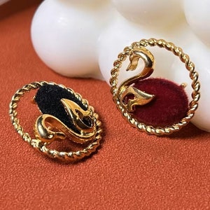 Pode incluir: Um par de brincos de cisne em tom dourado com um design de corda trançada. Os cisnes estão virados em direções opostas e são colocados sobre um fundo de veludo preto e vermelho.