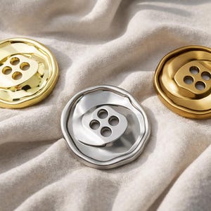 Puede incluir: Tres botones decorativos en tonos dorados y plateados. Cada botón tiene un diseño de cuatro agujeros y una forma orgánica texturizada. Los botones están dispuestos sobre una tela suave de color blanquecino, creando una composición simple pero elegante.