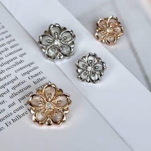 Könnte beinhalten: Drei blütenförmige Broschen mit Strasssteinen in der Mitte. Die Broschen sind silber, gold und roségold.