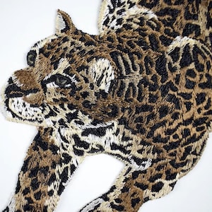 May include: Embroidered leopard patch. The patch features a detailed leopard design in shades of brown, black, and cream. The intricate stitching brings the animal to life. The leopard is shown in a dynamic pose, with its body partially visible.