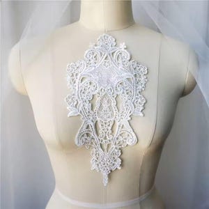 May include: White lace applique with a delicate floral design. The applique is shaped like a teardrop and has a scalloped edge.