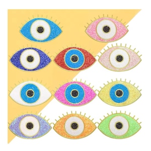 Könnte beinhalten: Satz mit zehn bunten bösen Augen-Anhängern mit Glitzerakzenten. Jeder Anhänger zeigt ein blaues Auge mit einer weißen Pupille und einer goldenen Umrandung. Die Anhänger sind in verschiedenen Farben, darunter Pink, Rot, Blau, Grün, Gelb und Lila.