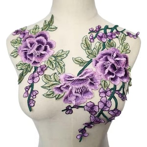 Op de afbeelding: Paarse bloemenborduursel applique met groene bladeren en wijnstokken. De applique heeft drie grote bloemen en kleinere bloemen langs de wijnstokken.