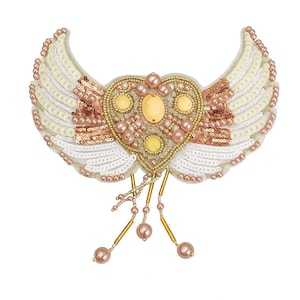 May include: A handcrafted, heart-shaped brooch with wing-like embellishments. The brooch features a central heart adorned with yellow and pink gemstones, surrounded by gold beads and sequins. The wings are white with rows of sequins and pearl accents.
