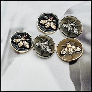 May include: Four round black and gold bee-shaped buttons with a black background and gold accents. The buttons have a 3D effect and are perfect for adding a touch of whimsy to any project.