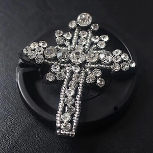 Op de afbeelding: Een zilveren, kruisvormige versiering met een grote, heldere strasssteen in het midden en kleinere strassstenen eromheen.