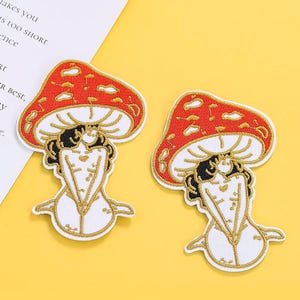 Könnte beinhalten: Zwei gestickte Bügelbilder mit einem roten und weißen Pilz, in dem eine Frau sitzt. Die Frau trägt ein schwarzes Oberteil und hat lange schwarze Haare. Die Pilze sind mit goldfarbenem Garn umrandet.