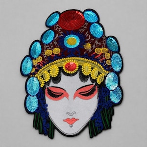 May include: A decorative embroidered patch featuring a stylized face with closed eyes, red eyeshadow, and red lips. The headpiece is adorned with bright blue sequined oval accents, a central red sequin circle, and yellow embroidered details.