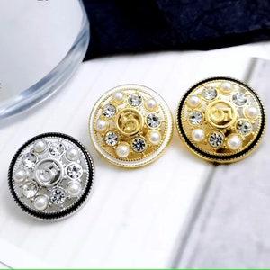 Puede incluir: Tres botones redondos decorativos con acabado dorado, plateado y negro. Cada bot&oacute;n presenta un n&uacute;mero "5" en relieve rodeado de diamantes de imitaci&oacute;n transparentes y cuentas blancas perladas.