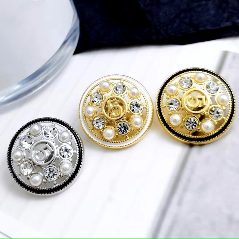 Puede incluir: Tres botones redondos decorativos con acabado dorado, plateado y negro. Cada bot&oacute;n presenta un n&uacute;mero "5" en relieve rodeado de diamantes de imitaci&oacute;n transparentes y cuentas blancas perladas.