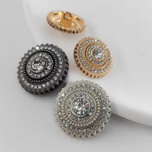 Puede incluir: Cuatro botones decorativos de forma redonda con un centro elevado. Los botones son de metal y tienen un acabado brillante. Los botones están adornados con diamantes de imitación y tienen una variedad de colores, incluyendo oro, plata y negro.