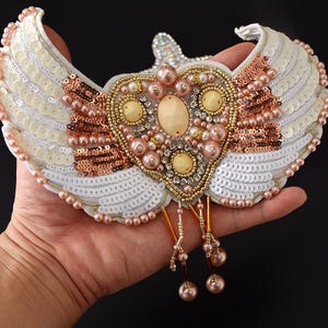 Op de afbeelding: Een decoratief, hartvormig ornament met witte paillettenvleugels en een centraal ontwerp van parels, kralen en edelstenen. De vleugels zijn afgezet met roze parels en het hart heeft een grote, ovaalvormige steen. Het ornament heeft gouden en koperen accenten.