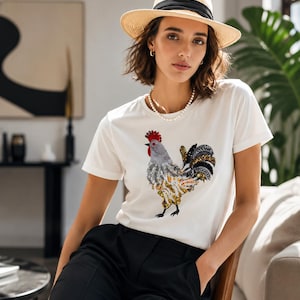 Pode incluir: Camiseta branca com um desenho de galo com lantejoulas vermelhas, prateadas, douradas e pretas. A pessoa está usando um chapéu de palha com uma faixa preta e calças pretas. Um colar de pérolas e brincos completam o look.