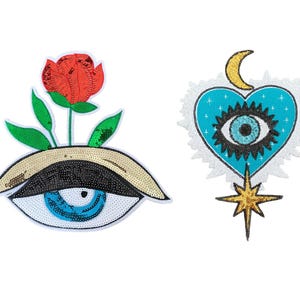 Könnte beinhalten: Zwei Pailletten-Patches auf weißem Hintergrund. Einer ist ein Auge mit einer roten Rose und grünen Blättern. Der andere ist ein blaues Herz mit einem Auge, einem goldenen Halbmond und einem goldenen Stern.