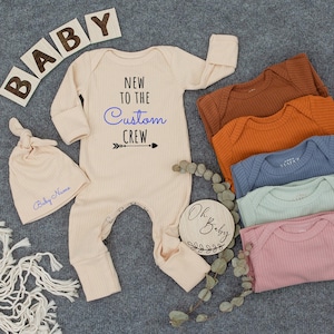 May include: A cream-colored baby onesie with the text "NEW TO THE CUSTOM CREW" and a matching knotted hat. Several other colored onesies are stacked nearby. Wooden blocks spell out "BABY". A wooden circle says "Oh Baby".