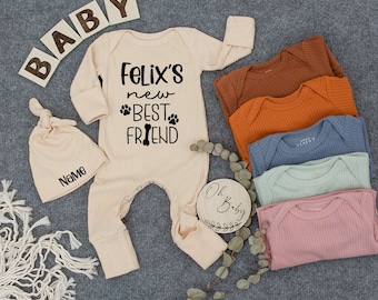 May include: A beige baby outfit set with a matching knotted hat. The outfit says "Felix's new BEST FRIEND" with paw prints and a bone graphic. Additional colored onesies in brown, orange, blue, green, and pink are also displayed.