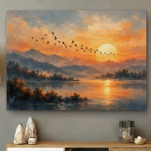 May include: A rectangular wall art print depicting a sunset over a calm lake with silhouetted mountains in the background. A flock of birds flies across the orange and yellow sky toward a large, pale sun. The water reflects the warm light.