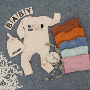 May include: An assortment of baby clothing and accessories in neutral tones. Includes a cream-colored onesie with the text "MILK, THEN BURPEES," a matching knotted hat with "BABY NAME," and a wooden sign that says "Oh Baby." Also, there are several ribbed bodysuits in various colors.