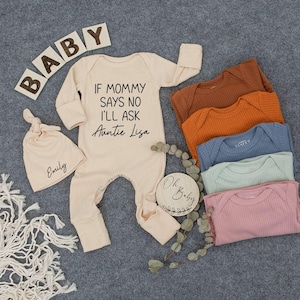 May include: A beige baby onesie with the text "If Mommy Says No I'll Ask Auntie Lisa". The onesie is next to a beige knitted baby hat with the name "Emily" embroidered on it. There are also several beige, orange, and blue ribbed baby onesies in the background.