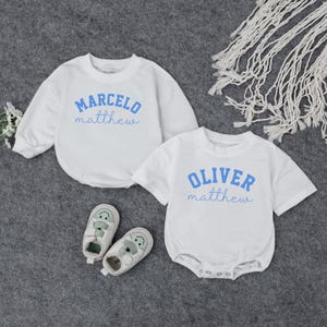May include: Two white baby rompers with blue text. One long-sleeved romper says "MARCELO matthew", the other short-sleeved romper says "OLIVER matthew". Two pairs of white and green baby shoes with smiling faces are also in the image.