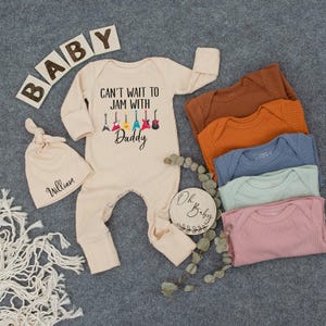 May include: A beige baby onesie with the text "Can't wait to jam with Daddy" and a row of colorful guitars. A beige knitted baby hat with the name "William" is also included. The onesie is surrounded by a variety of beige, brown, orange, blue, and pink ribbed baby onesies.