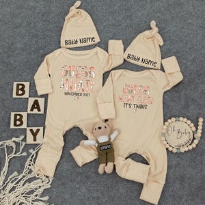 May include: Two beige baby onesies and matching hats with the text "GUESS WHAT?" and "GUESS WHAT ELSE? IT'S TWINS". The onesies have long sleeves and ribbed texture. A small teddy bear and wooden blocks spelling "BABY" are also present.