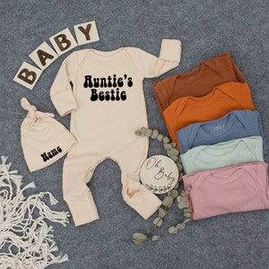 May include: A cream-colored baby onesie with the text "Auntie's Bestie" in black. The onesie is laid out on a gray surface with a wooden sign that says "Oh Baby" and a cream-colored knitted hat with the word "Name" on it.
