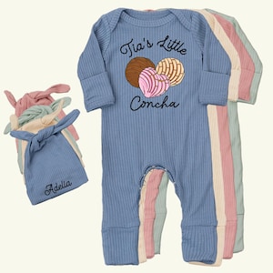 May include: Blue ribbed baby onesie with the text "Tia's Little Concha" and a graphic of conchas. Several matching knotted beanies in blue, pink, and cream are also visible. One beanie is personalized with the name "Adelia".