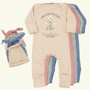 May include: A beige ribbed baby onesie with the text "Hand Picked for Earth Custom in Heaven" and a graphic of an elephant with balloons. Several matching pastel-colored baby hats are also visible.
