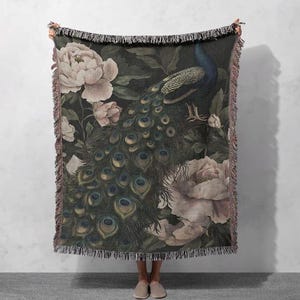 Peut inclure: Un plaid tissé présentant un fond sombre avec un grand paon déployant ses plumes de queue irisées, entouré de grandes fleurs de pivoine rose pâle. La couverture est dotée d'un bord à franges décoratif sur le haut et le bas.