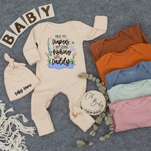 May include: A beige baby onesie with the text "Pack my diapers I am going fishing with Daddy" and a matching knotted hat. Also shown are several solid-colored baby bodysuits in various colors, and wooden blocks that spell "BABY".