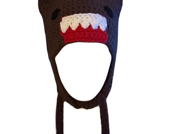 Pode incluir: Um chapéu castanho de crochê com um rosto de desenho animado. O chapéu apresenta uma boca branca com dentes vermelhos, olhos pretos e protuberâncias semelhantes a orelhas. Possui uma grande abertura para o rosto e duas peças penduradas.
