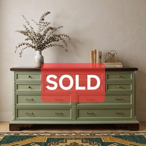 May include: A sage green dresser featuring a dark wood top and base, with eight drawers accented by small brass knobs and pulls. A white vase with dried eucalyptus branches, three books, and a gold wire sculpture sit on the top surface.
